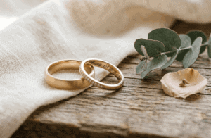 „Endlich tue ich etwas mit Wert“ – Interview mit Traurednerin Gabriele Mertesdorf 12 Zwei goldene Eheringe liegen auf einem Holztablett neben Eukalyptusblättern und einer Blüte, symbolisieren Liebe und Partnerschaft. Perfekt für Hochzeits- oder Verlobungsbilder.